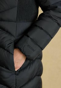 Melna pūkaina jaka ar quilted dizainu, vertikālām ribinātām paneļām, sānu kabatām un reljefu "Joules" logo uz piedurknes.