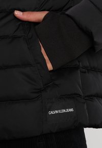 Czarna kurtka puchowa o pikowanej strukturze, z naszywką z logo Calvin Klein Jeans. Zawiera mankiety w czarnym kolorze.