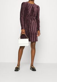 Mujer vestida con un vestido de terciopelo púrpura de manga larga y con relieve, sosteniendo un bolso blanco con una cadena dorada, y usando mocasines negros con acentos dorados.