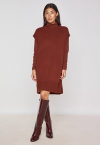 Abito lavorato a maglia marrone con collo alto e maniche corte, abbinato a stivali bordeaux al ginocchio, design minimalista e leggera trama a coste.