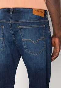 Tmavě modré džíny z denimu s koženou nášivkou označenou "DIESEL", dvěma zadními kapsami s charakteristickým žlutým prošitím a zúženým střihem.