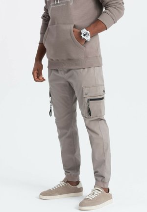 Pantalons cargo gris avec poches zippées, coupe fuselée et poignets élastiques, associés à des baskets gris clair et à un hoodie beige avec poche kangourou.