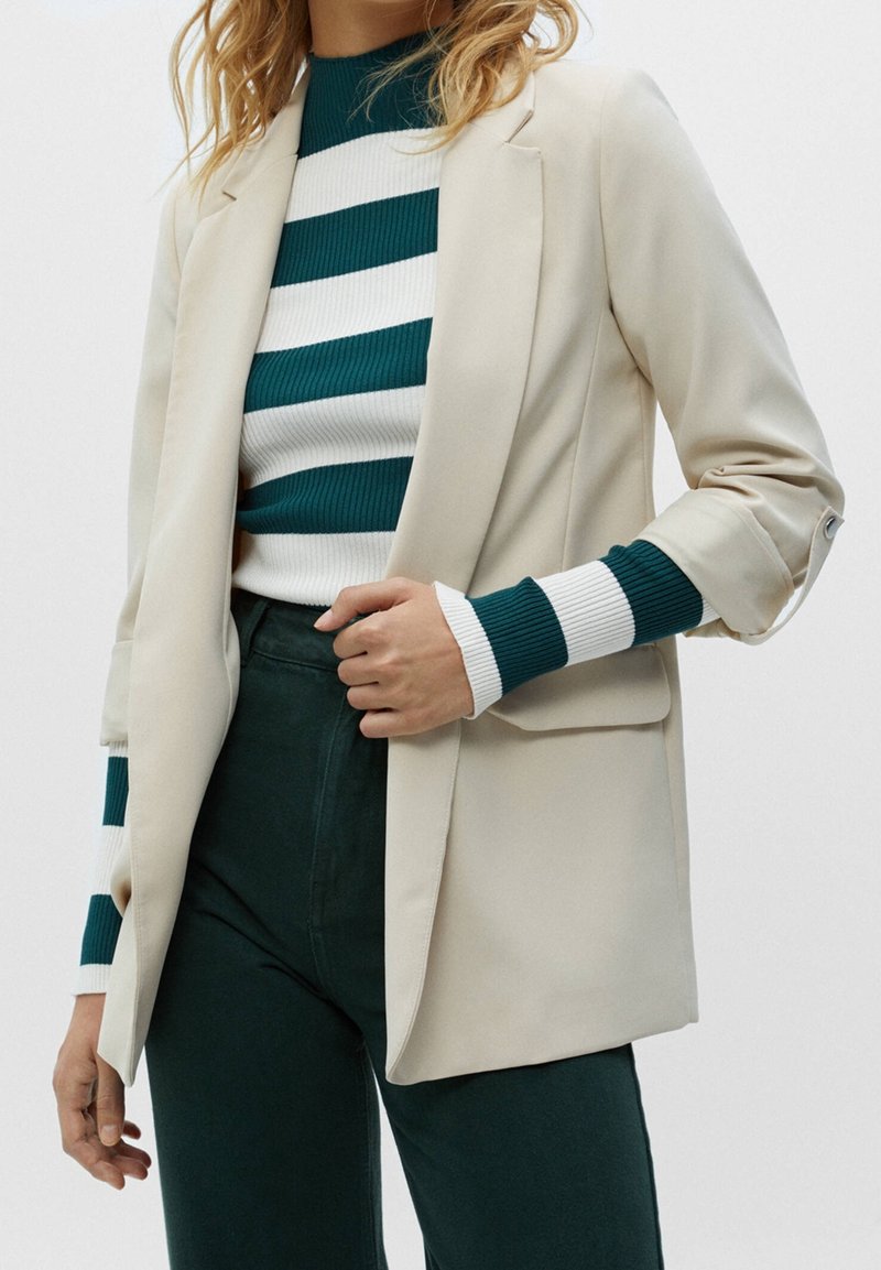 Femme portant un blazer beige sur un pull à manches longues rayé vert et blanc et un pantalon vert foncé, la main tenant le revers du blazer.