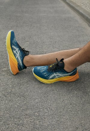 Beine mit blauen und gelben Laufschuhen ruhen auf grauem Asphalt nahe eines Bordsteins.