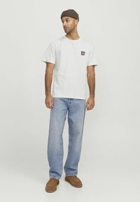 Hvid bomulds T-shirt med sort og rød grafisk tekst på brystet, parret med lyseblå denim jeans og brune slip-on sko.