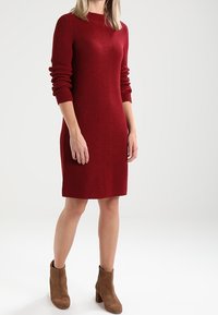 Rotes geripptes Strickkleid mit langen Ärmeln, hochgeschlossen und mit seitlichen Schlitzen, kombiniert mit braunen Stiefeln bis zum Knöchel.