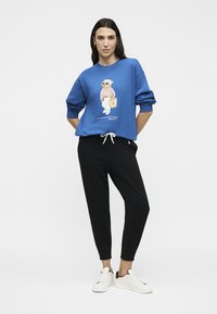 Γυναίκα με μπλε φούτερ με σχέδιο αρκούδας, μαύρο παντελόνι με κορδόνι και λευκά αθλητικά παπούτσια, στέκεται μπροστά από ένα απλό φόντο.