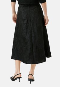 Jupe midi noire avec un motif floral embossé, coupe trapèze, fermeture éclair dissimulée, et texture lisse. Portée avec des sandales à talons noires.