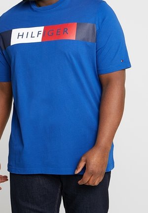 Mężczyzna w niebieskiej koszulce Tommy Hilfiger z czerwono-biało-granatowym logo na piersi i ciemnych spodniach na jednolitym tle.