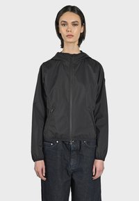 Giacca nera leggera con cappuccio, chiusura con zip e tasche laterali. Texture liscia e polsini elasticizzati, abbinata a jeans scuri larghi.