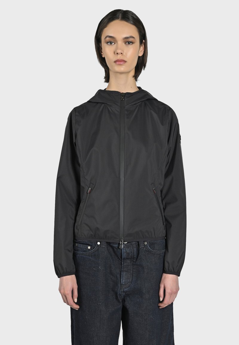Giacca nera leggera con cappuccio, chiusura con zip e tasche laterali. Texture liscia e polsini elasticizzati, abbinata a jeans scuri larghi.