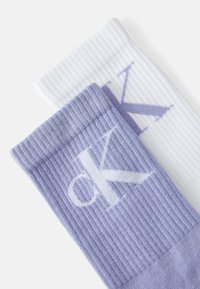 Chaussettes blanches et lavande côtelées arborant un logo brodé bien visible. Le design comprend des couleurs contrastées et des bandes texturées.