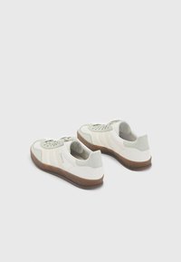 Pár off-white tenisek Adidas Gazelle se světle šedými akcenty a gumovými podešvemi umístěnými na bílém pozadí.
