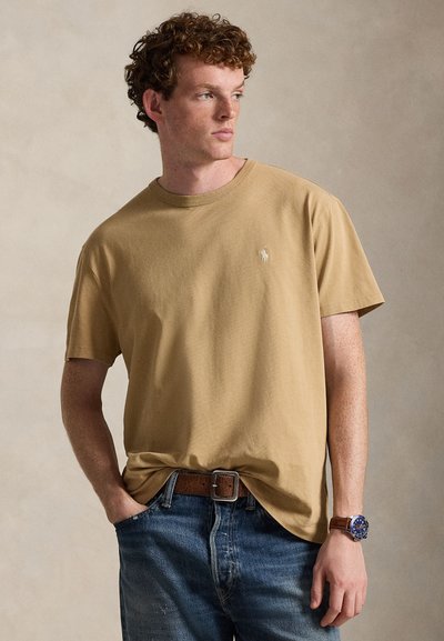 Tan cotton t-shirt with a round neckline and short sleeves, featuring a small embroidered logo on the chest. Jeans with a brown belt.