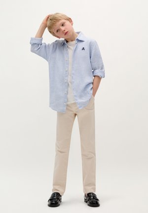 Chemise à rayures bleu clair avec un T-shirt blanc en dessous, pantalon beige et chaussures noires brillantes. La chemise comporte un petit logo brodé.