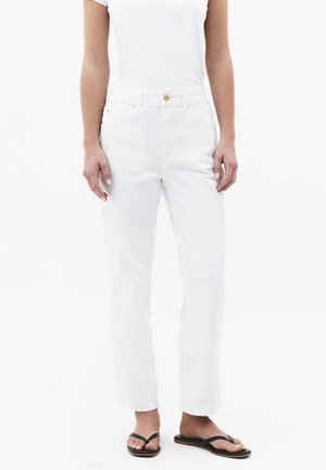 Person iført tætsiddende hvide jeans og en hvid kortærmet top kombineret med sorte flip-flops mod en hvid baggrund.