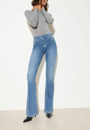 Kvinde iført lys grå langærmet top, højtaljede blå flare jeans og sorte spidse hæle står mod hvid baggrund.