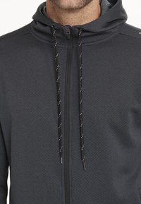 Harmaa vetoketjuhuppari, jossa on teksturoitu kangas, musta vetoketju ja kuviolliset nyörit. Ominaisuuksiin kuuluu korkea kaulus ja istuva muotoilu.