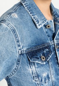 Denim jacket in faded light blue, featuring distressed details, two chest pockets, silver-tone button closures, and a classic collar design.