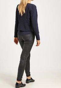 Blazer bleu marine à coupe ajustée, dos superposé, associé à un pantalon en cuir noir et des chaussures plates noires, offrant un look épuré et moderne.