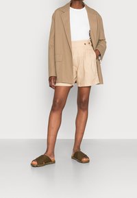 Beżowa marynarka z jedną kieszenią, biała koszulka, kremowe szorty z guzikami oraz brązowe sandały na krzyżujących się paskach z teksturowaną podeszwą.