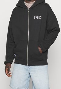 Mies, który ma na sobie czarną bluzę z suwakiem z napisem "PEQUS" na klatce piersiowej i jasnoniebieskie dżinsowe szorty, stoi z jedną ręką w kieszeni.