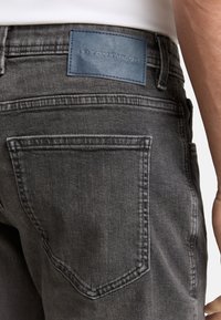 Dark gray denim jeans with a leather brand patch, detailed stitching, and a classic five-pocket design. Close-up of the back view.