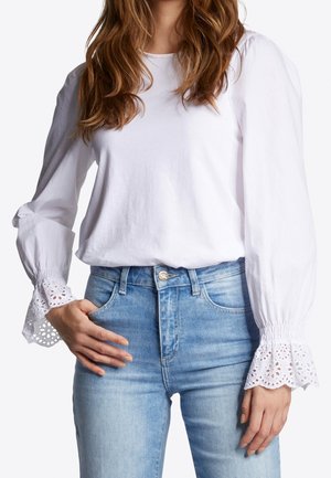 Vrouw draagt een witte blouse met lange mouwen met opengewerkte kanten manchetten en lichtblauwe high-waisted jeans, hand rust in zak.