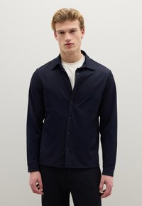 Camicia blu navy con bottoni, colletto morbido, maniche lunghe, taschino singolo sul petto e tessuto liscio e leggero. Indossata sopra una maglietta bianca a girocollo.