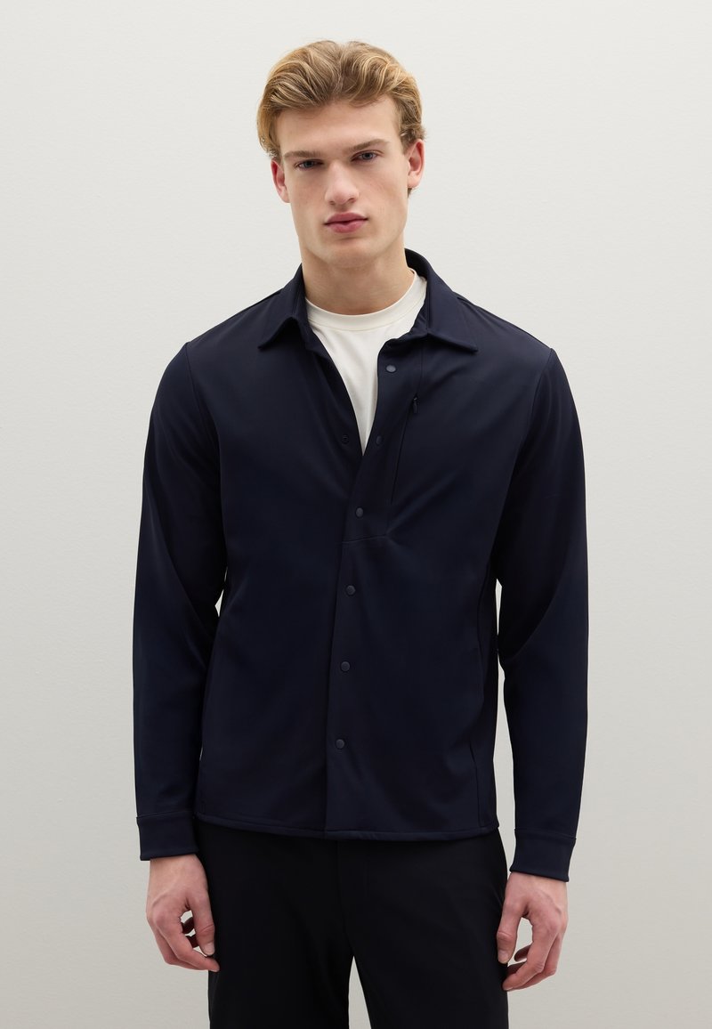 Camicia blu navy con bottoni, colletto morbido, maniche lunghe, taschino singolo sul petto e tessuto liscio e leggero. Indossata sopra una maglietta bianca a girocollo.