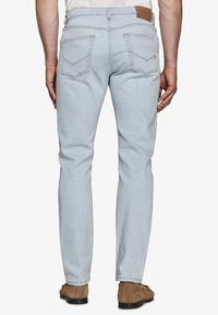 Hombre con jeans ajustados de color azul claro y zapatos de ante marrones, de pie con los brazos relajados a los lados, de espaldas.
