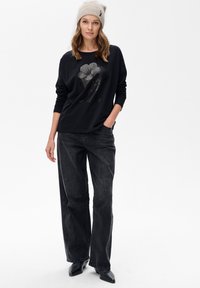 Czarna koszula z długim rękawem z motywem kwiatowym, w połączeniu z luźnymi ciemnymi dżinsami. Model nosi jasnoszary wełniany beret i czarne buty.