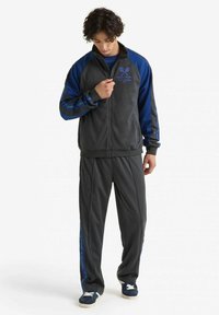Completo sportivo grigio e blu con giacca zip-up dalle maniche a contrasto, cuciture decorative e pantaloni abbinati con strisce laterali.