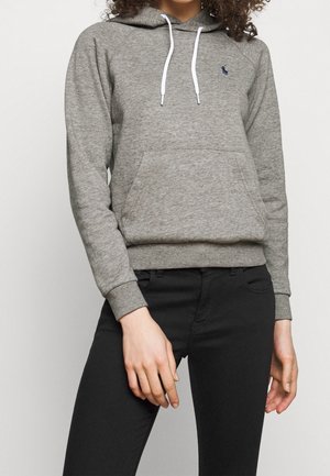 Vrouw die een grijze capuchonsweater met witte trekkoorden en een zwarte broek draagt, staand met ontspannen handen langs haar kanten.