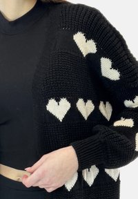 Zwarte gebreide cardigan met witte hartpatronen, met een relaxte pasvorm en geribbelde manchetten. Gedragen over een zwart top, die het ontwerp benadrukt.