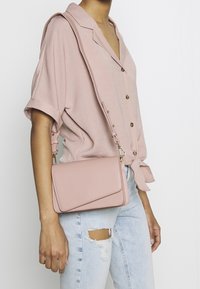 Světle růžová kabelka přes rameno vyrobená z hladkého materiálu. Má klopu na zavírání a nastavitelný popruh. Nosená s ležérním tričkem a roztrhnutými džíny.
