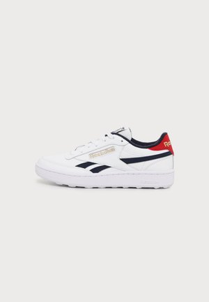 Baskets blanches Reebok avec des bandes noires, une languette rouge au talon, un logo doré et une semelle blanche, présentées sur un fond uni gris clair.