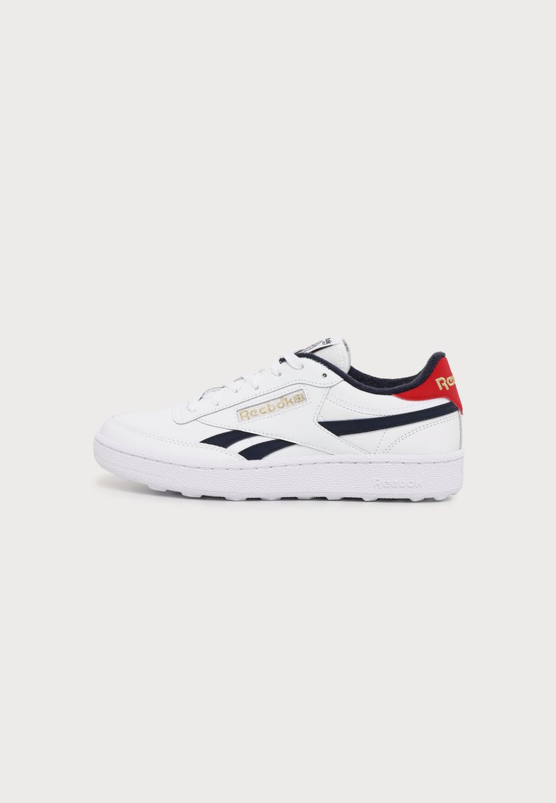 Λευκό αθλητικό παπούτσι Reebok με μαύρες ρίγες, κόκκινη γλωττίδα στη φτέρνα, χρυσό λογότυπο και λευκή σόλα, απεικονισμένο σε απλό ανοιχτό γκρι φόντο.