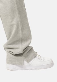 Weißer Sneaker mit perforierter Zehenkappe, Schnürdesign und strukturiertem Sohle. Getragen mit hellgrauen Jogginghosen, die eng anliegen und tapered Beinen haben.