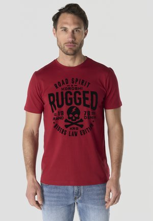 Hombre vestido con una camiseta roja con gráfico de calavera y huesos cruzados en negro y texto, combinado con vaqueros azul claro, de pie frente a un fondo gris.