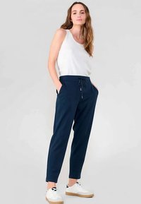 Marineblauwe taps toelopende broek met elastische tailleband en trekkoord, voorzien van zijzakken. Gecombineerd met een witte mouwloze top en witte sneakers.