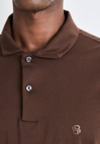 Brun polo-skjorte laget av mykt stoff, med krage, tre-knappestolpe og en liten logo på venstre bryst.