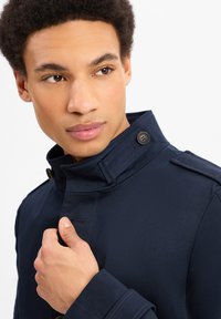 Navyblauwe trenchcoat met een hoge kraag, zichtbare stiksels en knopenaccenten. Materiaal lijkt glad en lichtjes waarop textuur te zien is.