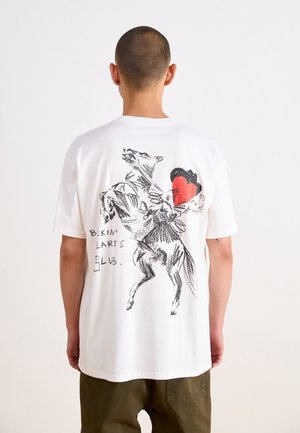 Person med kort hår bærer en hvid T-shirt med en sort skitse af en hest, et rødt hjerte og teksten "Bikini Hearts Club" på ryggen.