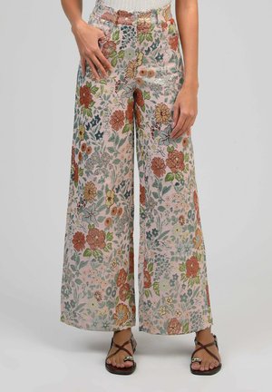 Pantalon à jambes larges avec motif floral multicolore, porté avec un haut blanc et des sandales à fines lanières foncées, mains reposant près des poches.