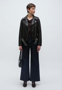 Μαύρο δερμάτινο μπουφάν μοτοσικλέτας, εφαρμοστό καφέ τοπ, τζιν με φαρδιές παντελόνες σε σκούρο χρώμα, καφέ μποτάκια αστραγάλου και μαύρη τσάντα χειρός. Λείες υφές και κλασικός σχεδιασμός.