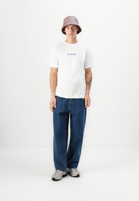 Modell trägt ein weißes T-Shirt mit einem bunten Grafikdesign, baggy blaue Jeans und einen helllila Bucket Hat. Graue Sneaker runden das Outfit ab.