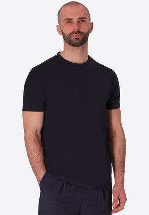 Uomo con la barba che indossa una semplice t-shirt nera a maniche corte e pantaloni scuri, in piedi con una mano in tasca su sfondo chiaro.