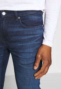 Vaqueros de mezclilla azul oscuro con un corte ajustado, que presentan bolsillos frontales, un cierre de botón y un ligero desvanecimiento. Acompañados de una camisa blanca de manga larga.