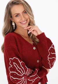 Červený vlněný kardigán s V výstřihem, stříbrnými knoflíky a zdobenými rukávy, které mají bílé kontury a červené flitry v květinových vzorech.